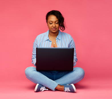 Apply Online Person sitting cross-legged using a laptop against a pink backdrop, wearing a blue striped shirt, jeans, and sneakers.