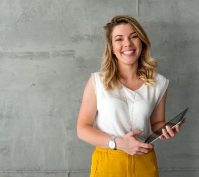 Candidate Presentation Smiling person stands against a concrete wall, holding a tablet, wearing a white top and mustard-yellow pants.