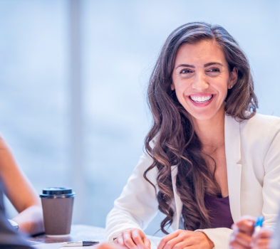 Candidate Screening & Selection Smiling professional in a white blazer sits at a meeting table with a coffee cup, hands poised, bright office backdrop.