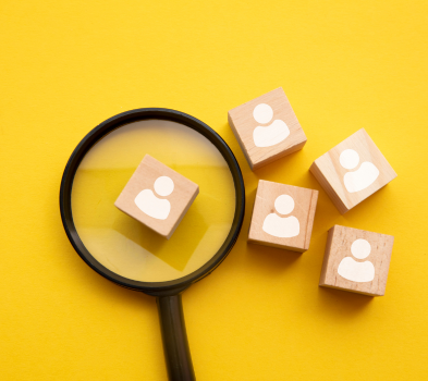 FOR EMPLOYERS Magnifying glass over five wooden blocks marked with person icons on a bright yellow background.