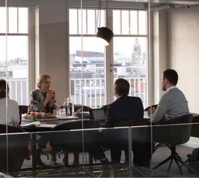 Interview with Priority Team meeting in a glass-walled conference room with city view; four colleagues seated around a table in discussion.