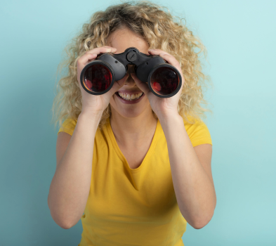Job seekers Curly-haired person in a yellow T-shirt smiles while looking through black binoculars against a light blue background.