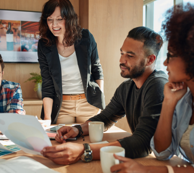 Local and Personal Team meeting around a table; a woman stands smiling and reviews a printout with colleagues holding coffee mugs.