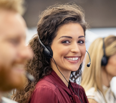 Ongoing Support Smiling call center agent wearing a headset looks at the camera, seated with teammates in a support office