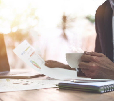 Role Profile & Sourcing Strategy Person reviews printed charts at a sunlit desk, holding a steaming coffee mug, with laptop and notebook nearby.