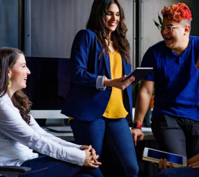 Skill & Review Group of coworkers chatting in an office; one standing holds a tablet while others seated nearby smile and laugh
