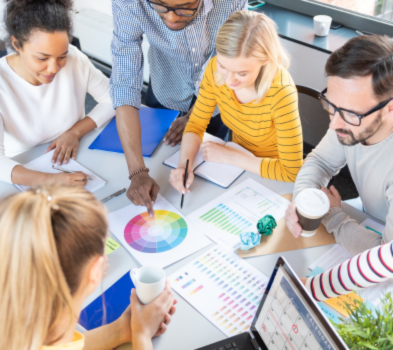 WHAT WE OFFER Team collaborating around a table, pointing to a color wheel and analytics charts during a design planning session.